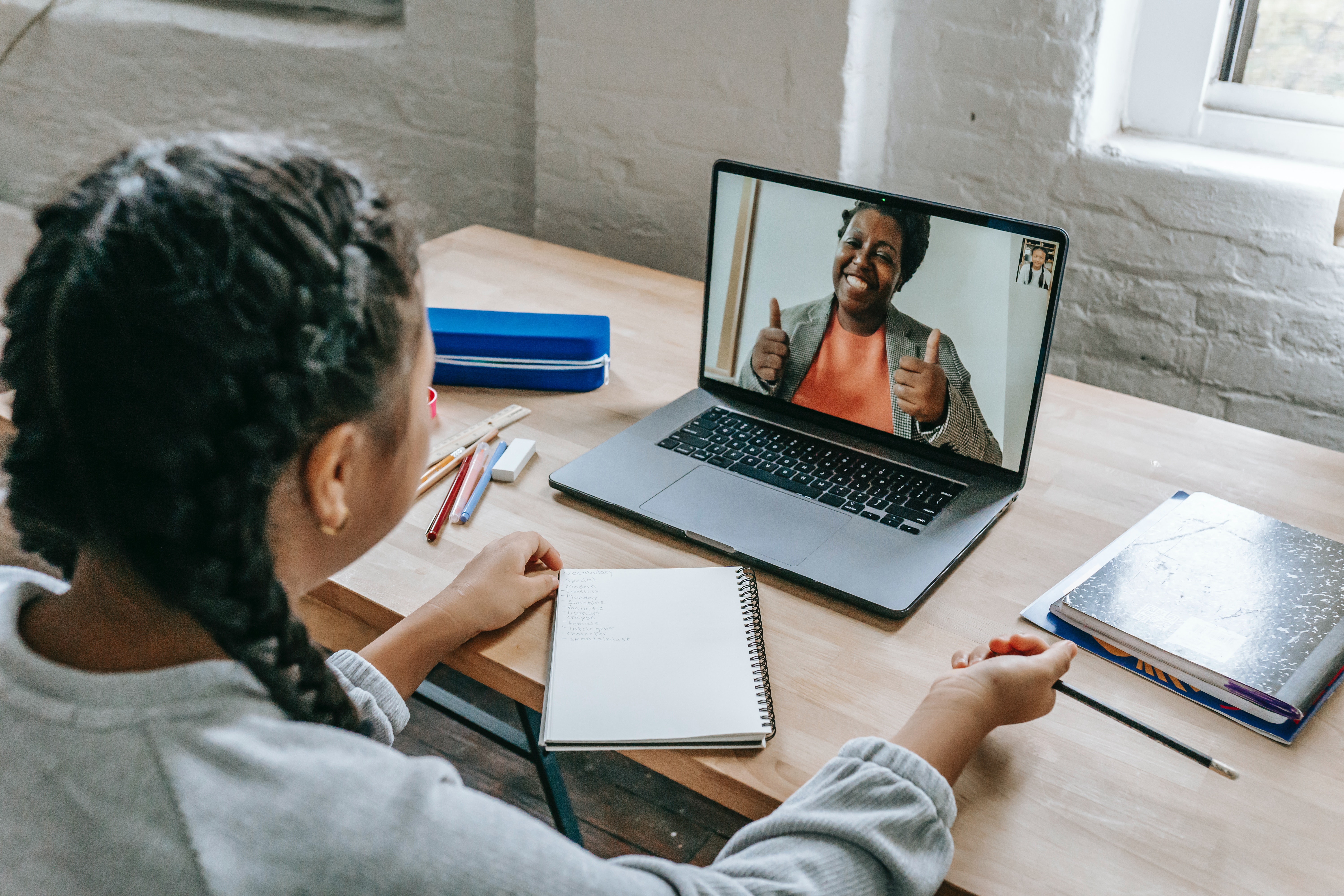 Woman sitting at her desk with a notebook and pencils, while collaborating with another woman on the computer screen who is smiling and giving two thumbs up.