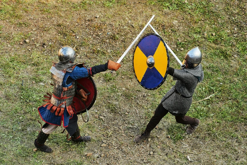 Two people in medieval warrior armor attack each other in mock combat with swords and large round shields.