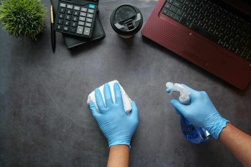 Person wearing blue cleaning glove, a spray bottle, and a cleaning towel in front of their laptop.