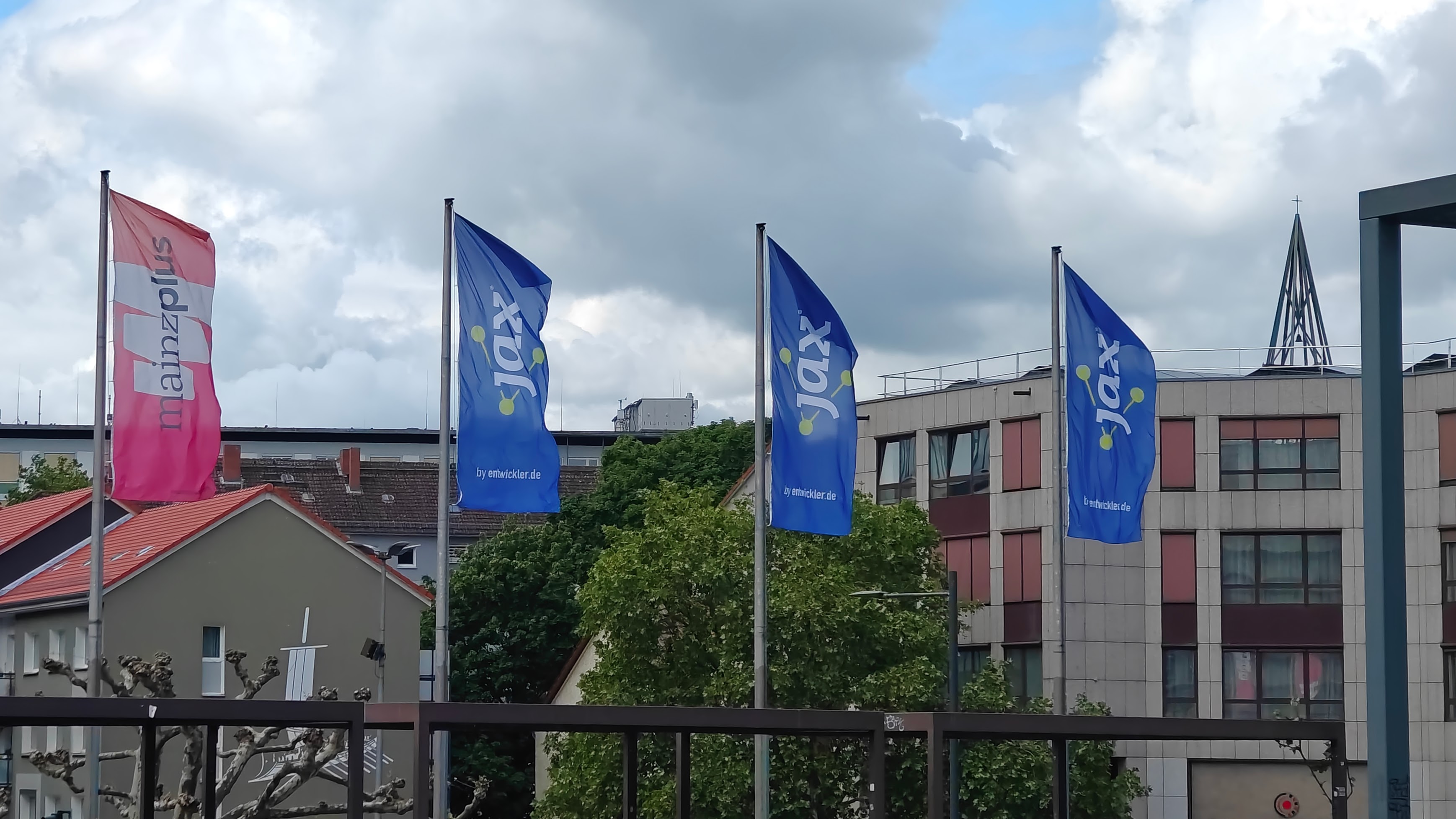 Photo of four vertical flags blowing in the wind. The leftmost flag is pink and white with the text "Mainz Plus" while the remaining flags are blue with white text, "JAX". The skyline has buildings up to the halfway point on the flags.