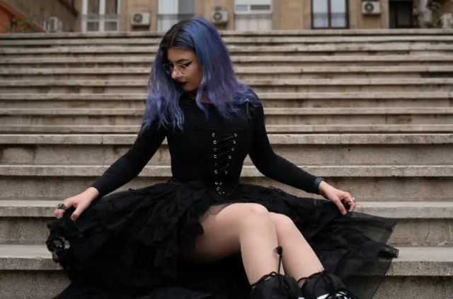 Woman with blue hair wearing black shoes and black dress sitting on some steps.