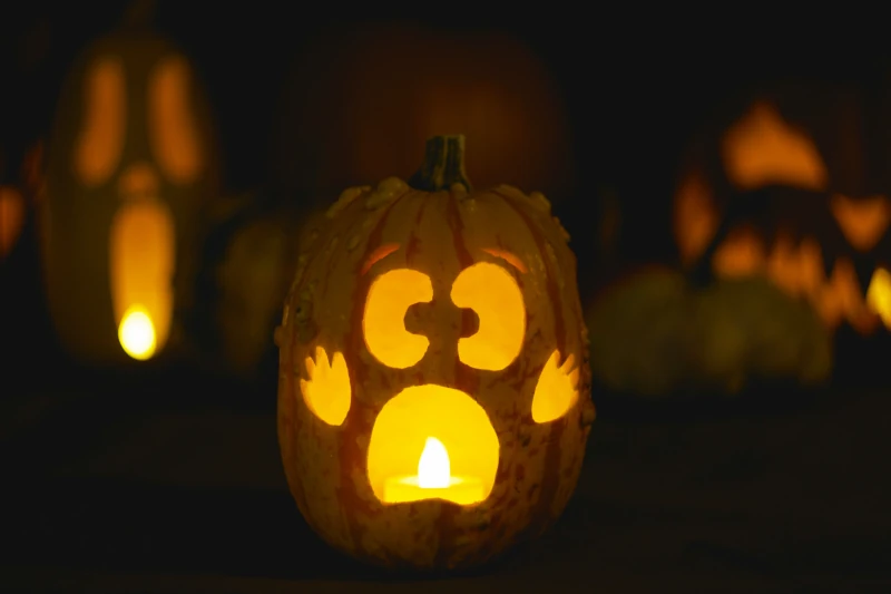 A candle lit carved jack-o-lantern. The carved face looks scared. Little hands are also carved on the sides of the pumpkin and the lantern looks like it is holding its hand up.