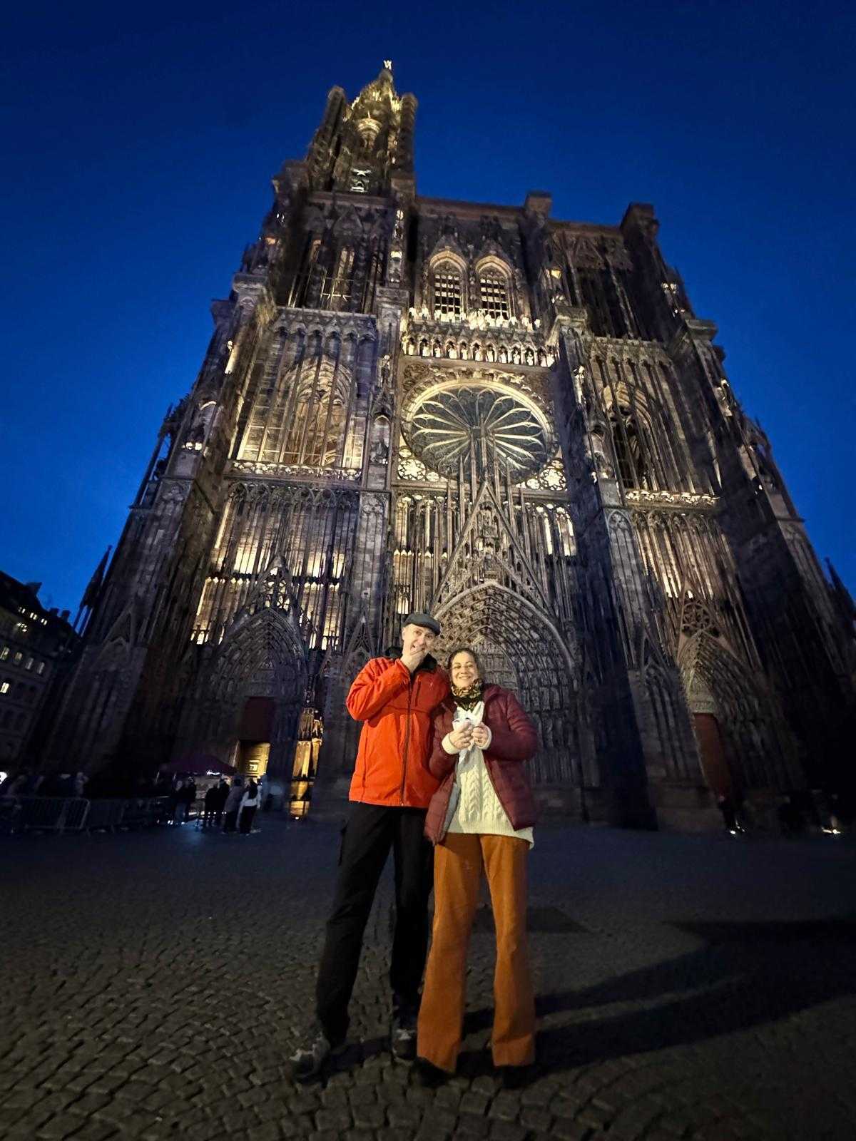 MartineMichaelCathedral Martine and Michael are standing in front of the Strasbourg Cathedral at night. The shot is from ground level, with the Cathedral towering over them.