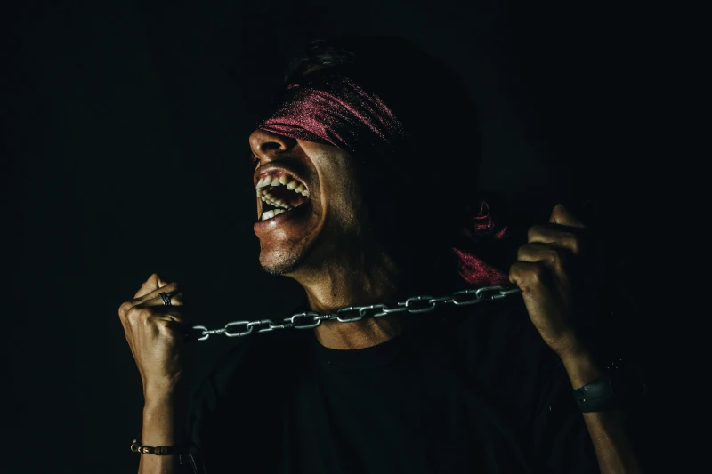 Screaming man wearing a red blindfold while straining to pull apart a chain between his hands.