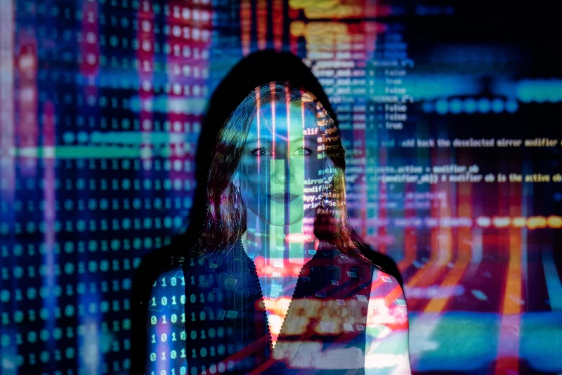 Woman standing against a wall as binary numbers and source code are projected against her and the wall in bright colors.