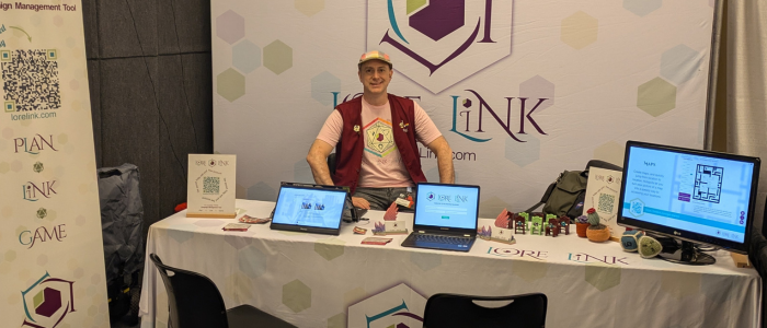 An image of the Lore Link booth at Gen Con. There are two chairs in front of a table that has monitors available for demos. There's a banner to the left with a QR Code sending people to get more info. There's a large banner in the back with the Lore Link name and logo, and there is a man sitting in front of the word Lore.