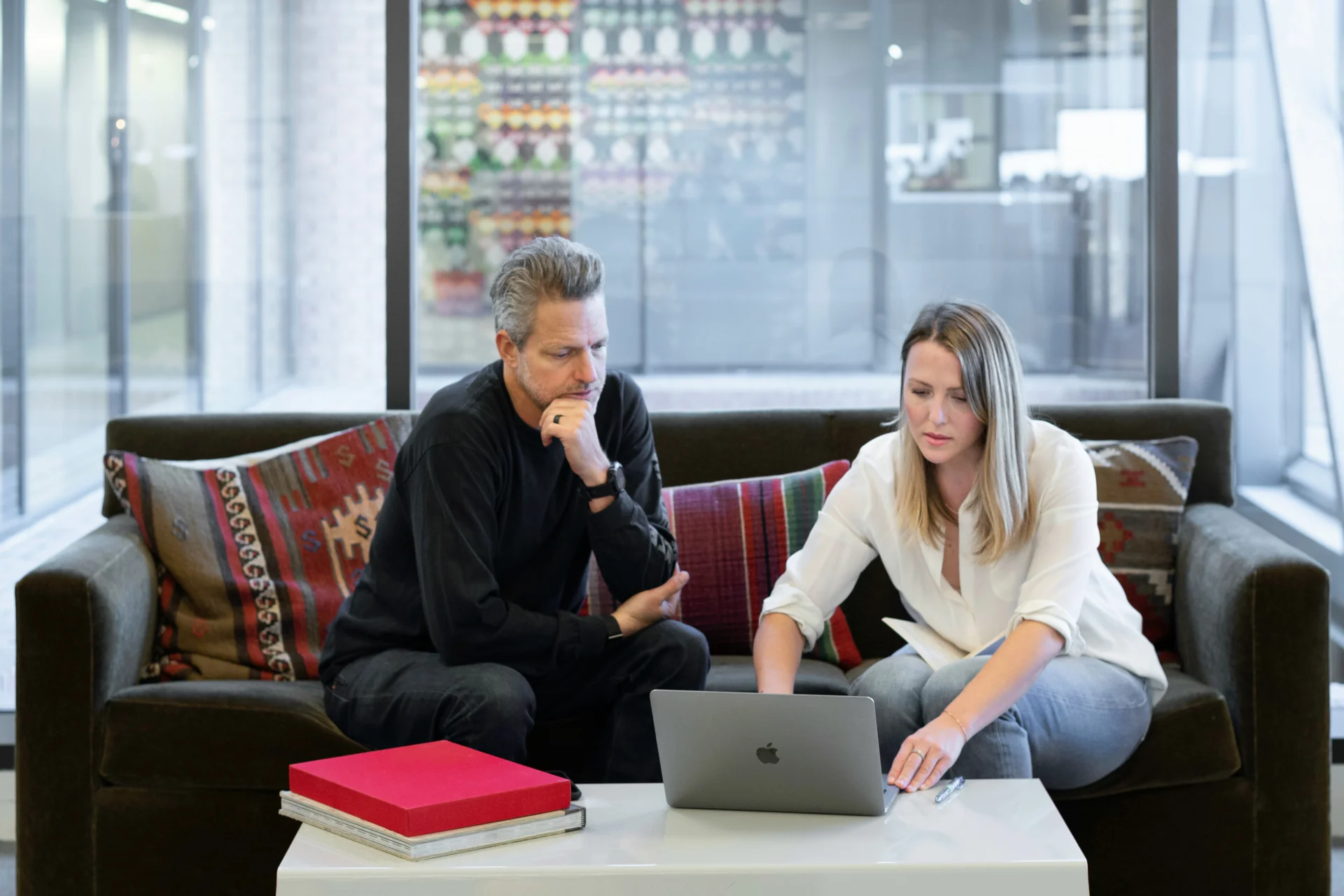 Two people sitting on a coach in a business meeting, looking at a laptop screen together.