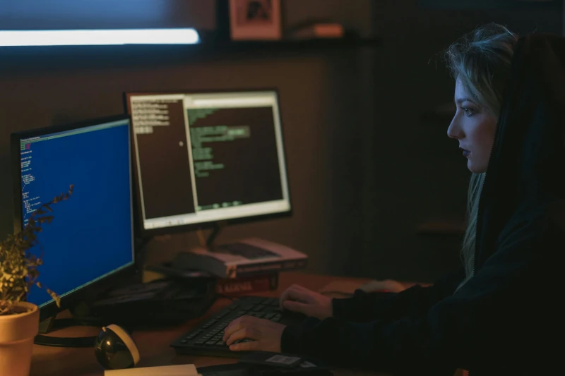 Woman in a dark room wearing a hoodie sitting at a desk coding at her computer.