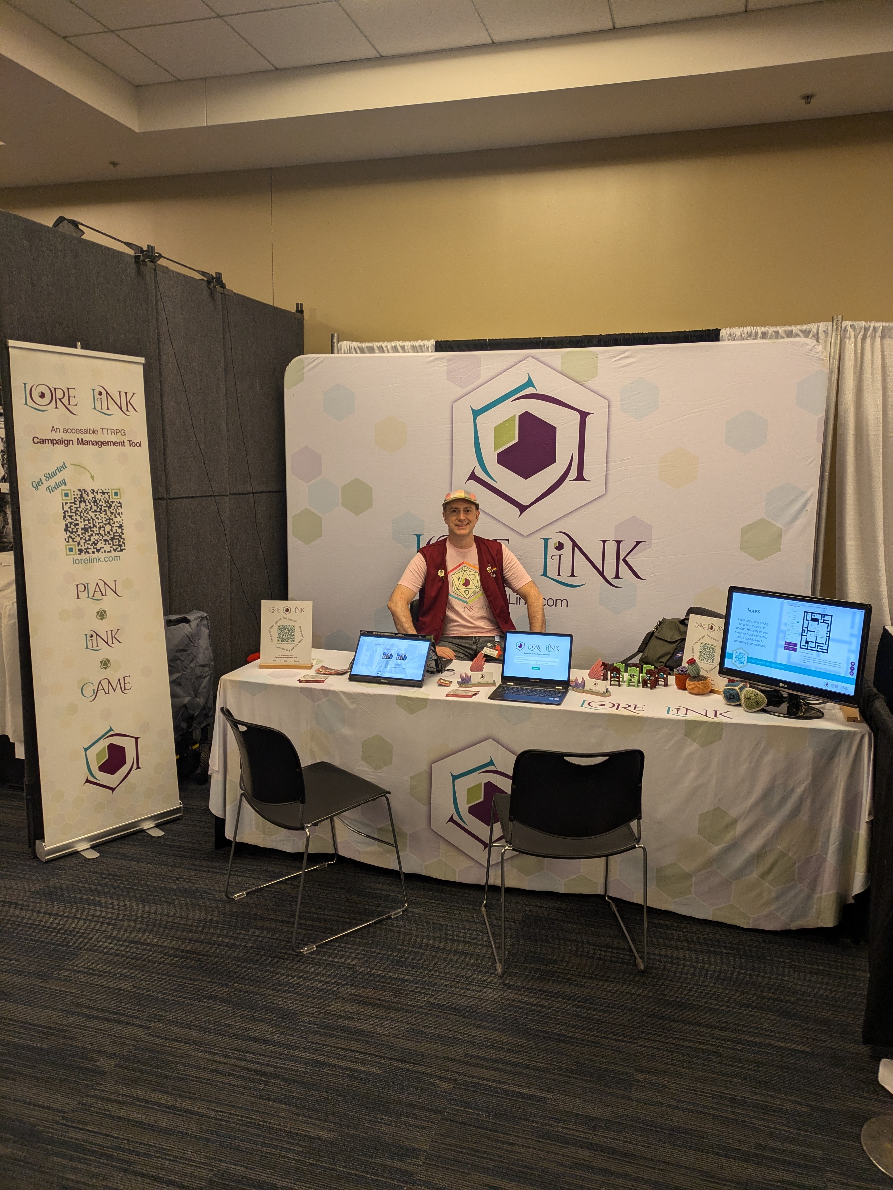 An image of the Lore Link booth at Gen Con. There are two chairs in front of a table that has monitors available for demos. There's a banner to the left with a QR Code sending people to get more info. There's a large banner in the back with the Lore Link name and logo, and there is a man sitting in front of the "lore."