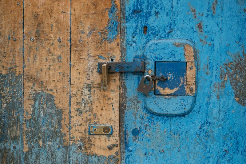 Very weathered double door, one with faded brown, peeling paint and the other with chipped light blue paint. There is a lock on the door and two padlocks fastening two different hasps on the doors.