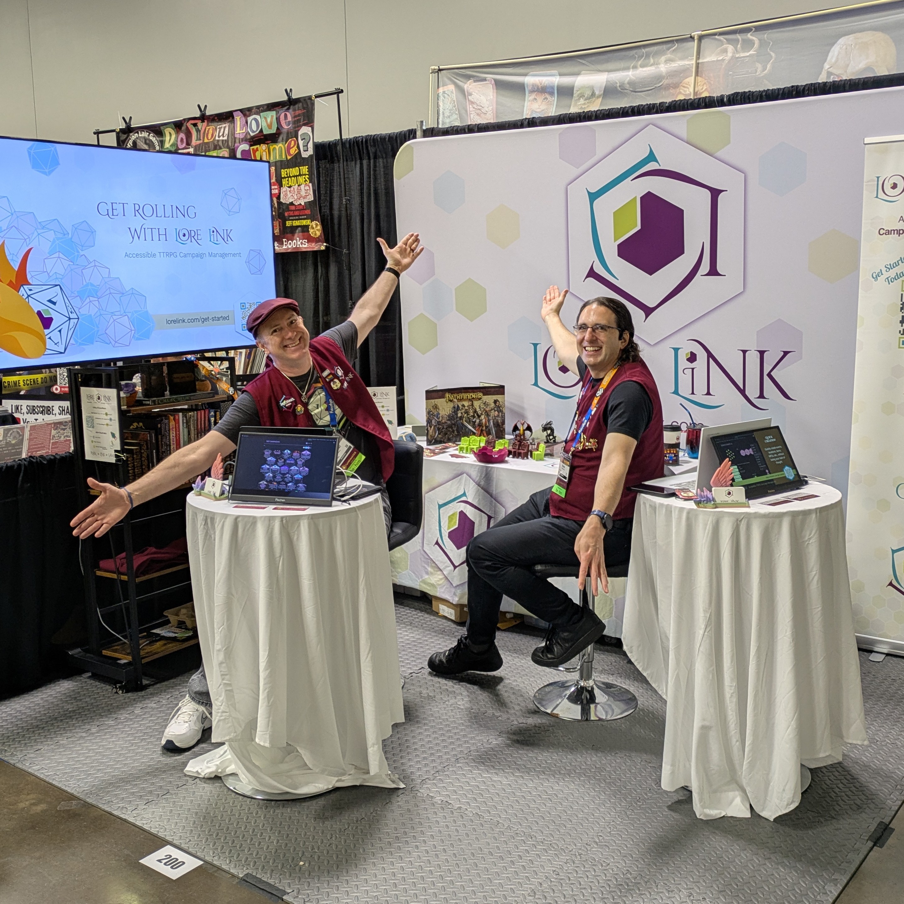 A photo of the Lore Link booth at Origins. It's a corner booth, with a large banner to the right and a large television to the left that reads Get Rolling with Lore Link. There are two small round tables with monitors for demos, and behind the tables on stools are Michael and Mark with their arms outstretched like they're going Ta Da!