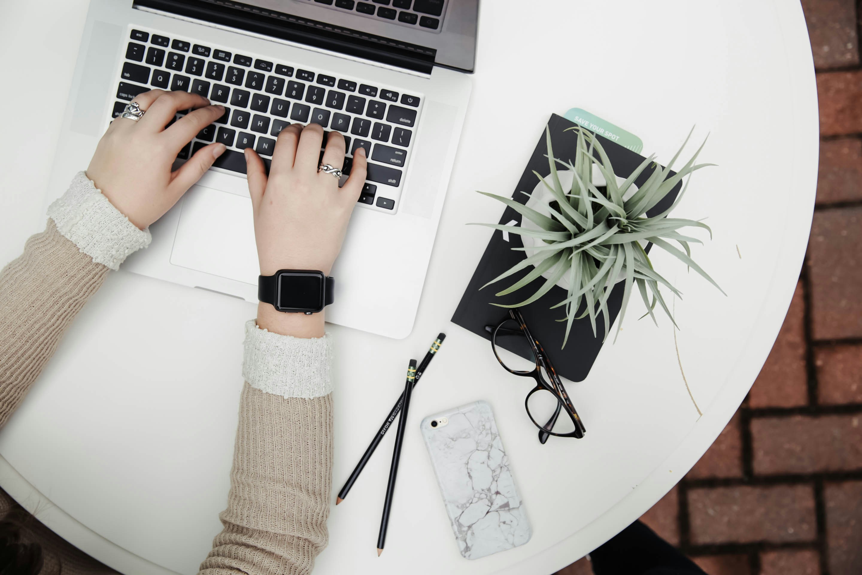 Top-down view of a person's hands with silver rings on each finger and a smart watch poised over a laptop keyboard sitting at a round white table. Next to them on the table is a green leafy plant sitting on top of a planter, a pair of black-rimmed glasses, two black pencils overlapping each other, and a face-down smartphone in a white and grey marble-texture case.