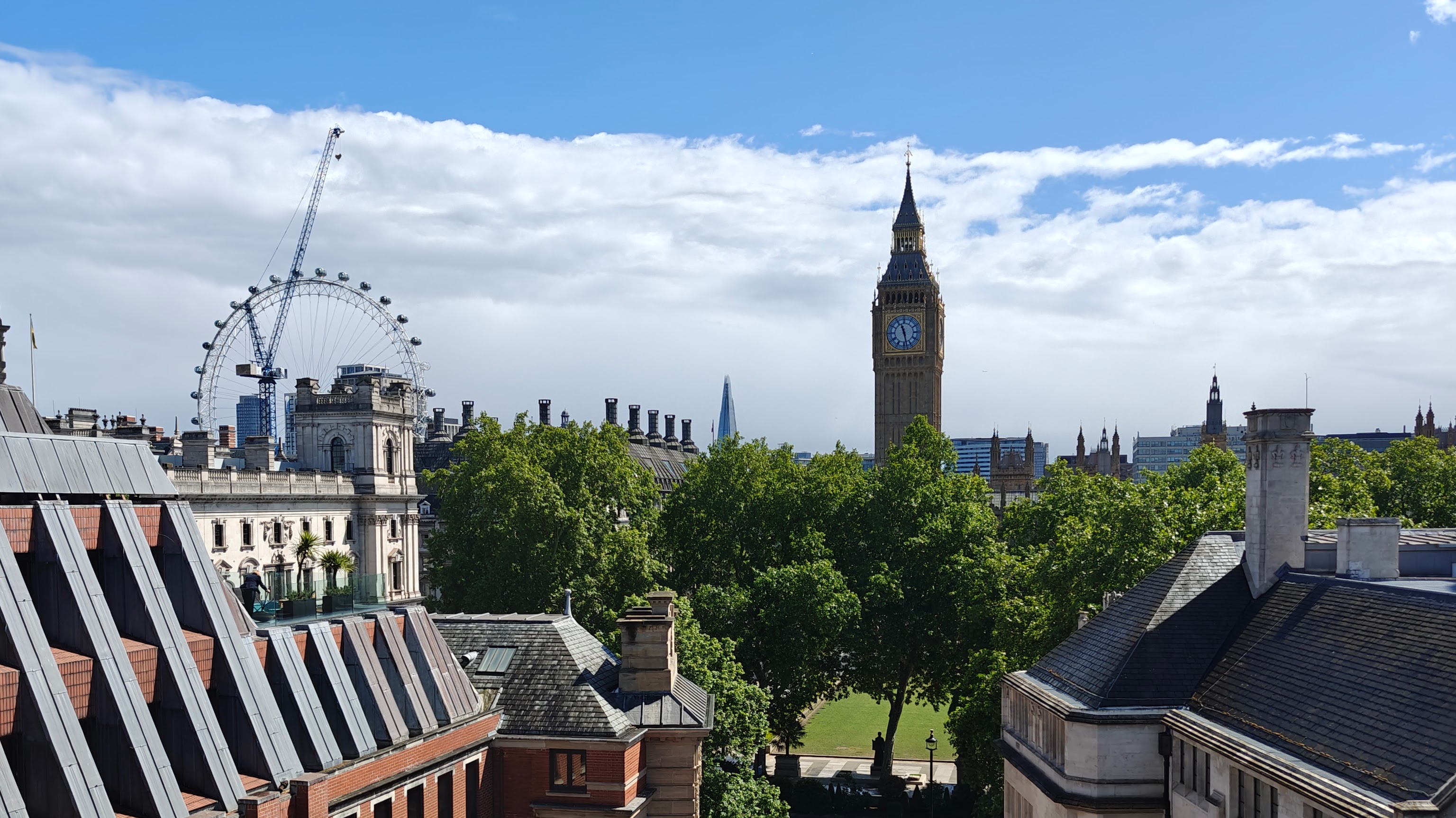 Photo of London featuring Big Ben in the center against a bright blue yet cloudy sky, with the London Eye to the left.