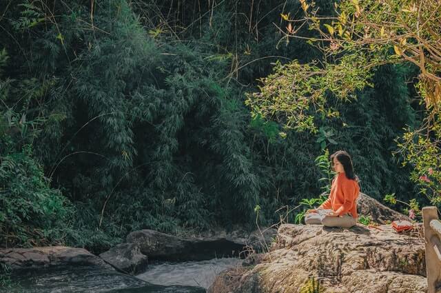 woman meditating next to river in forest