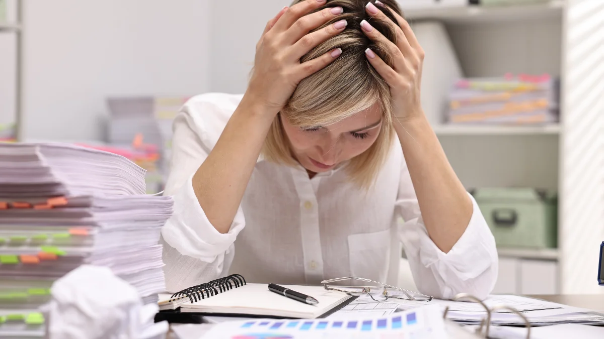 Woman sitting at her desk with her head in her hands, next to a stack of papers and a notebook.