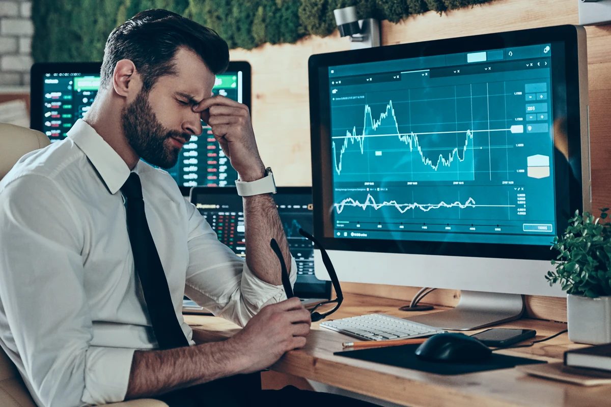 A frustrated man in a white shirt and tie sits at a desk with multiple monitors displaying financial graphs, suggesting stock market analysis or trading concerns.