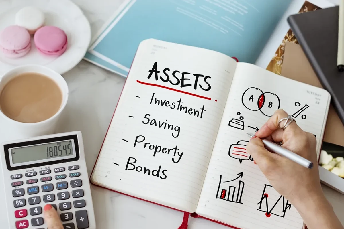 Open notebook with "Assets" written at the top listing investment, saving, property, and bonds, surrounded by a calculator, cup of coffee, macarons, and various charts.