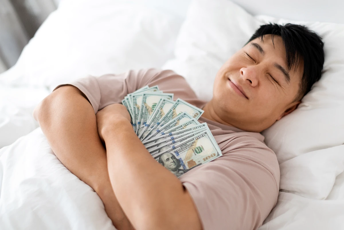 Man sleeping contentedly in bed while holding a fan of hundred-dollar bills against his chest.