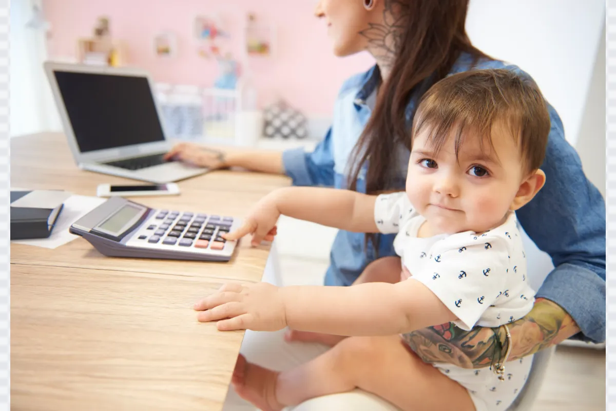 A baby sitting on their parent's lap at a desk, curiously pressing buttons on a calculator, while the parent works on a laptop.