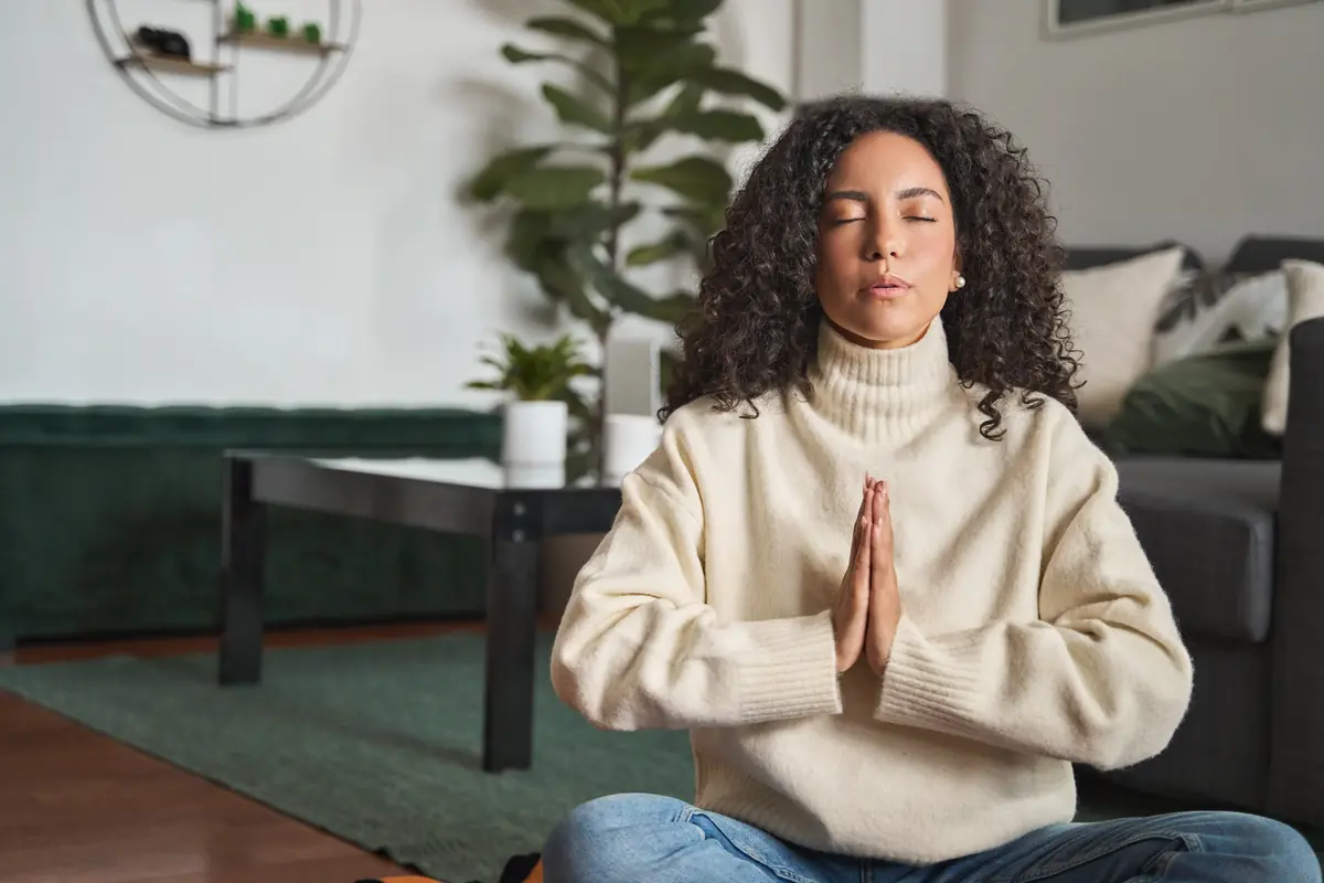 A person with closed eyes sits cross-legged in a meditative pose, wearing a cream turtleneck sweater, with their hands in a prayer position; a cozy living room setting with plants and a modern coffee table in the background.