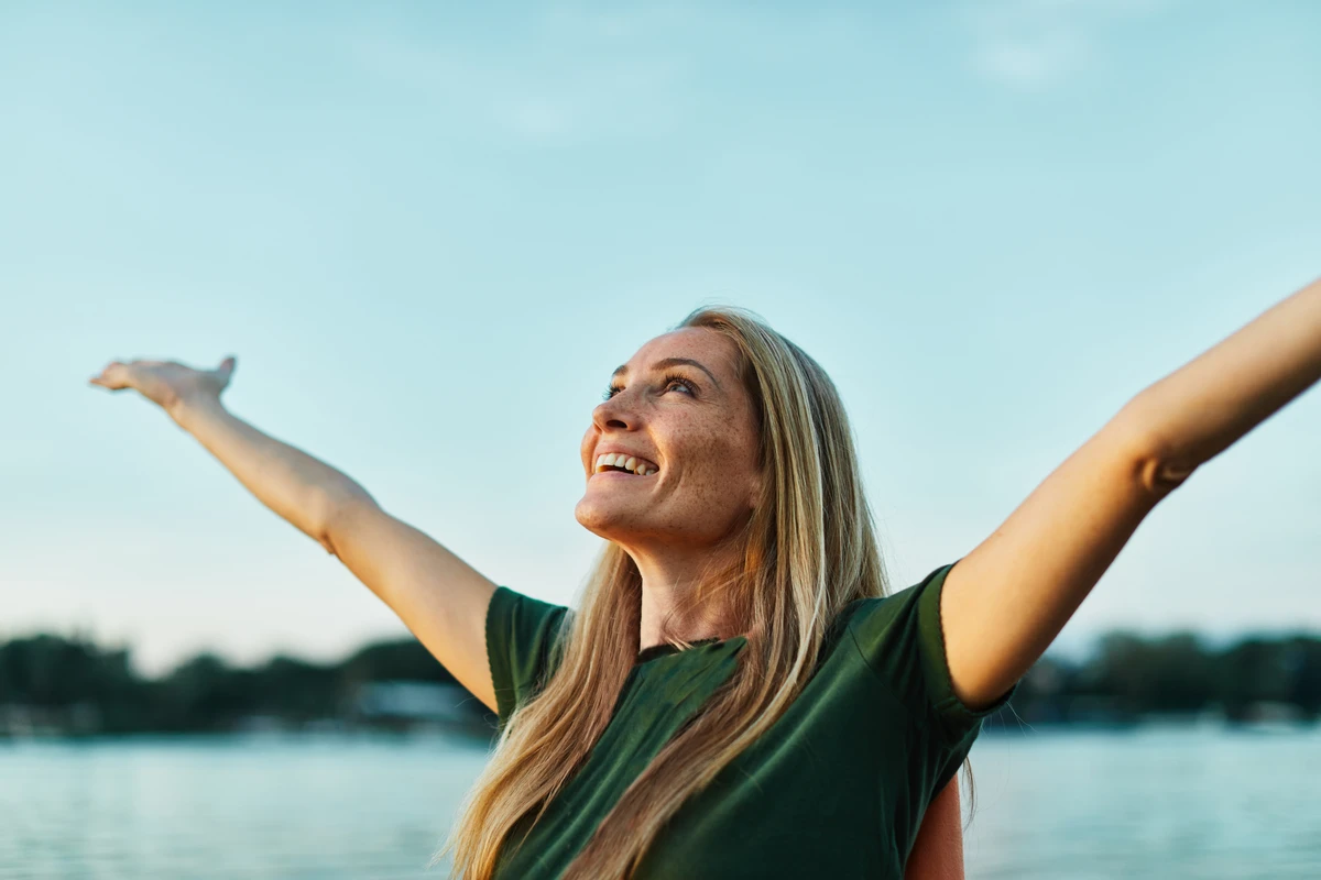 Woman smiling and spreading her arms wide against a backdrop of a clear blue sky and distant water.