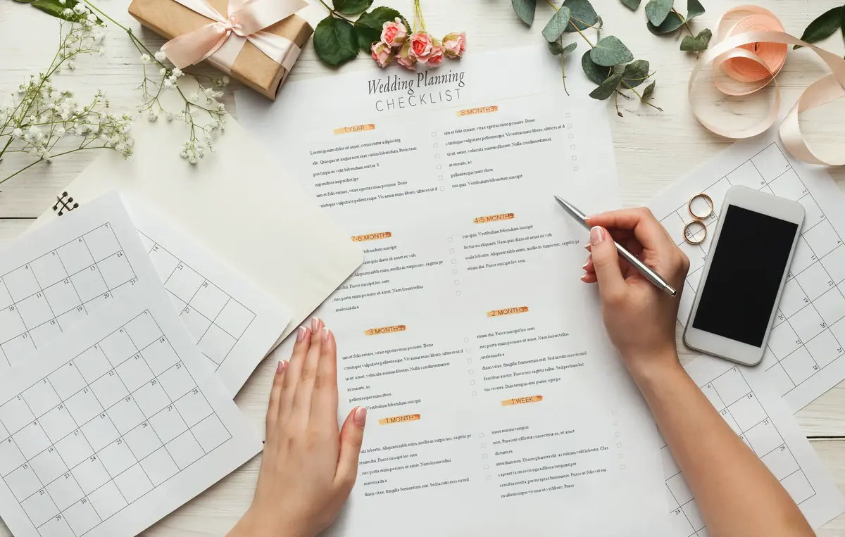 Hands working on a wedding planning checklist surrounded by a calendar, smartphone, flowers, rings, and gift box on a wooden surface.