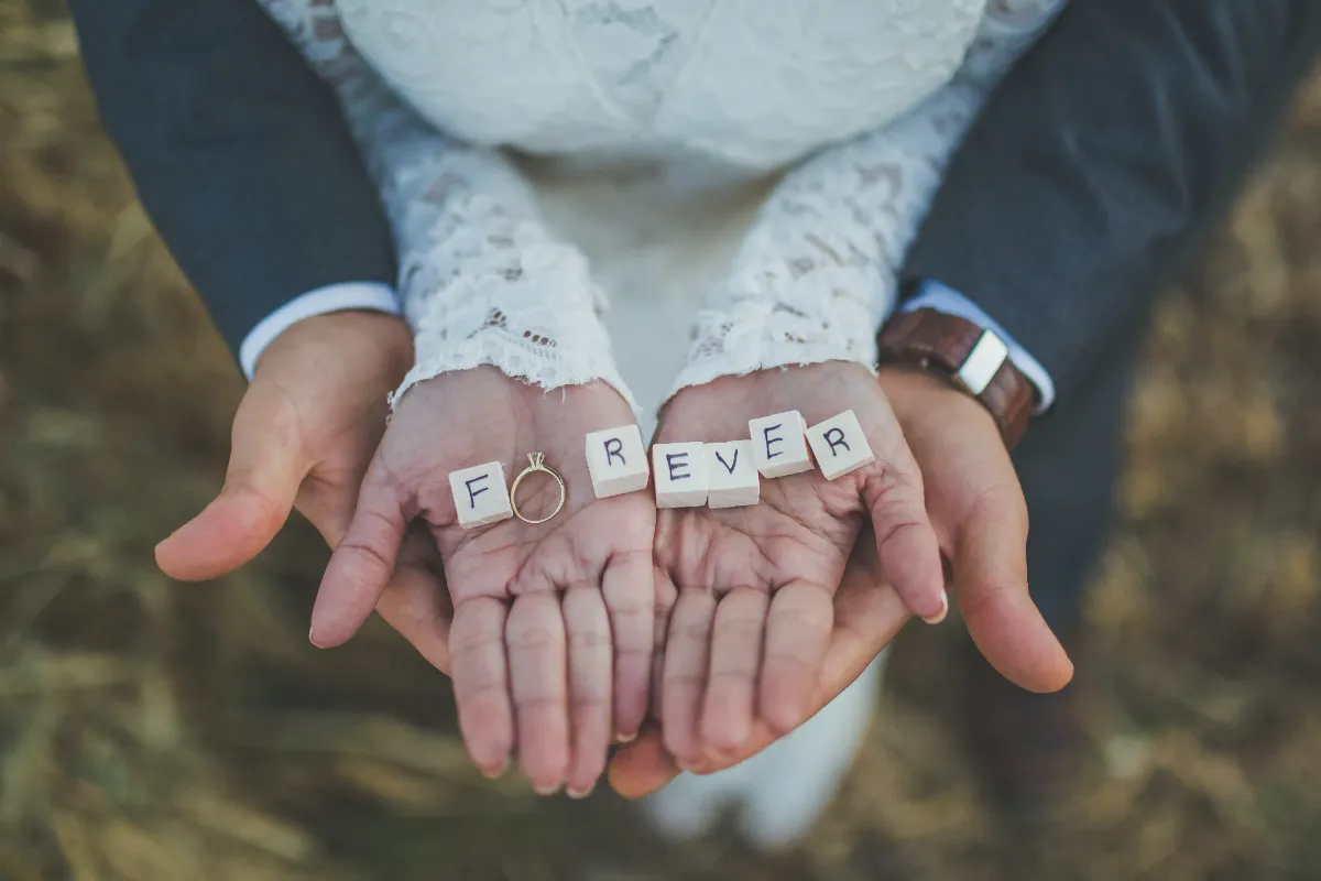 A bride and groom holding Scrabble tiles spelling "FOREVER" with a wedding ring resting on the bride's hand.