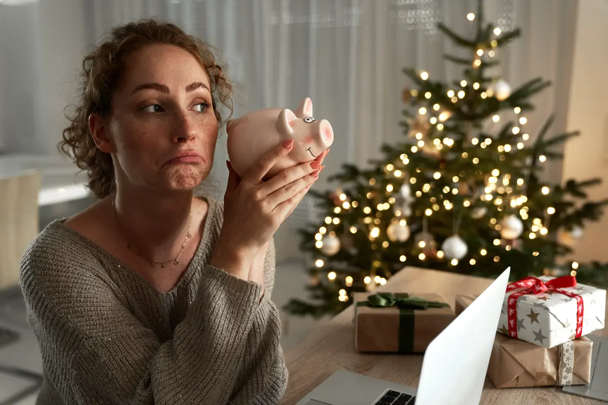 Woman looking at an empty piggy bank with a disappointed expression, sitting at a table with a laptop and Christmas tree in the background decorated with lights and ornaments.
