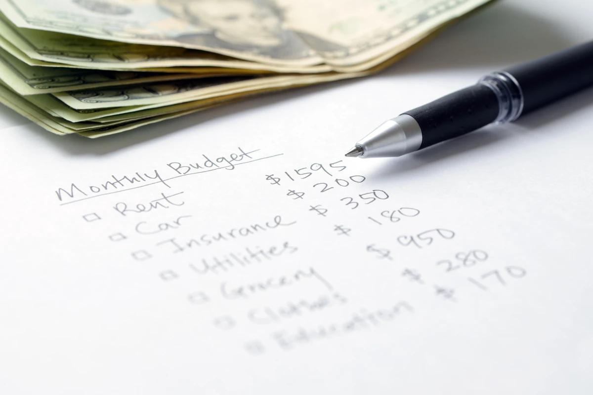 Handwritten monthly budget list on white paper with expenses for rent, car, insurance, utilities, grocery, and education next to a stack of dollar bills and a black pen.