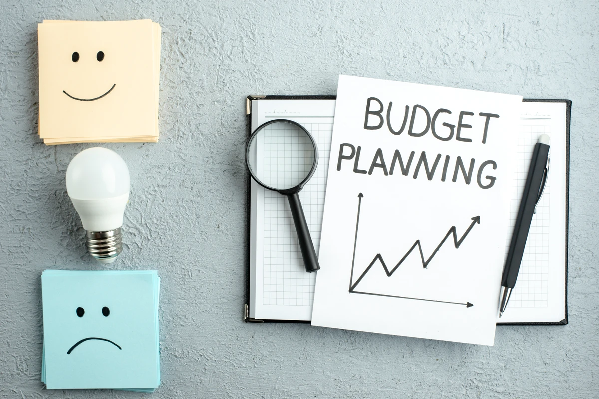 Sticky notes with smiley and frowny faces, a light bulb, magnifying glass, and pen surround a budget planning document featuring an upward trending chart on a textured surface.