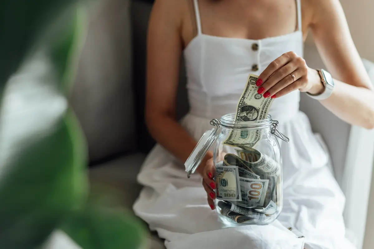 Woman in a white dress placing cash into a glass jar labeled for a budget wedding, with her nails painted red.