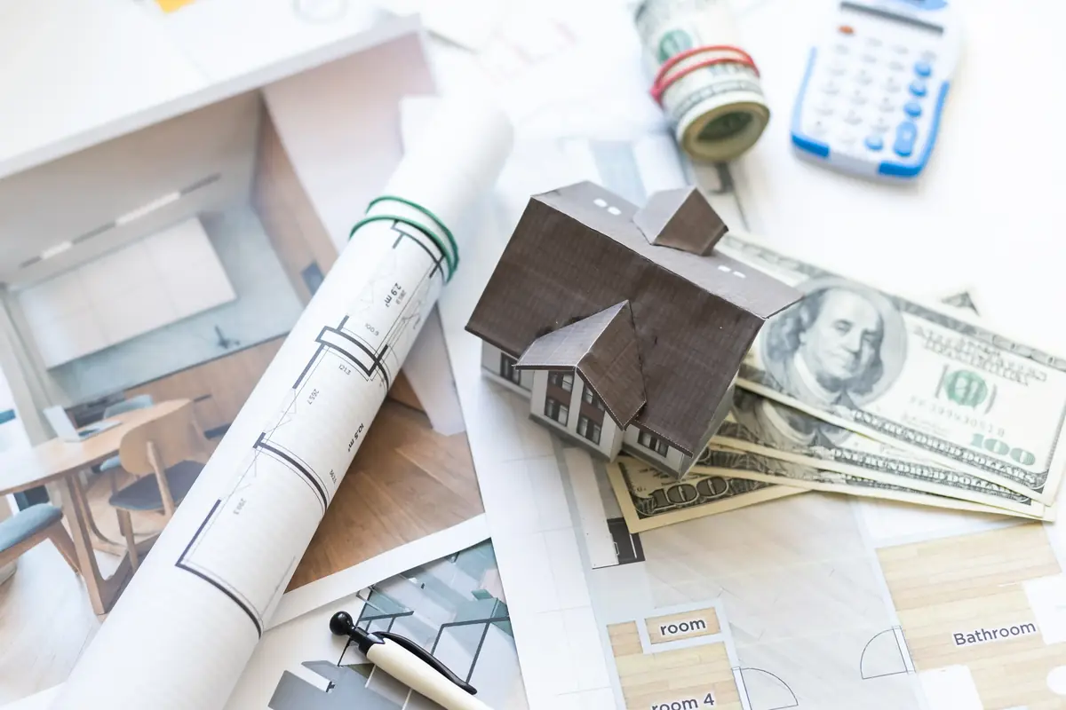 A small model house rests on architectural blueprints alongside a stack of hundred-dollar bills, a calculator, and a rolled-up blueprint.