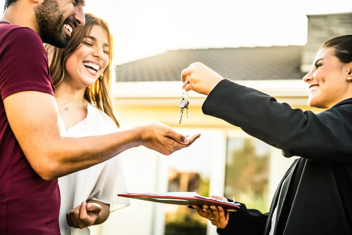 A couple joyfully receives keys from a professional, symbolizing the purchase of their new home, with a blurred house in the background.