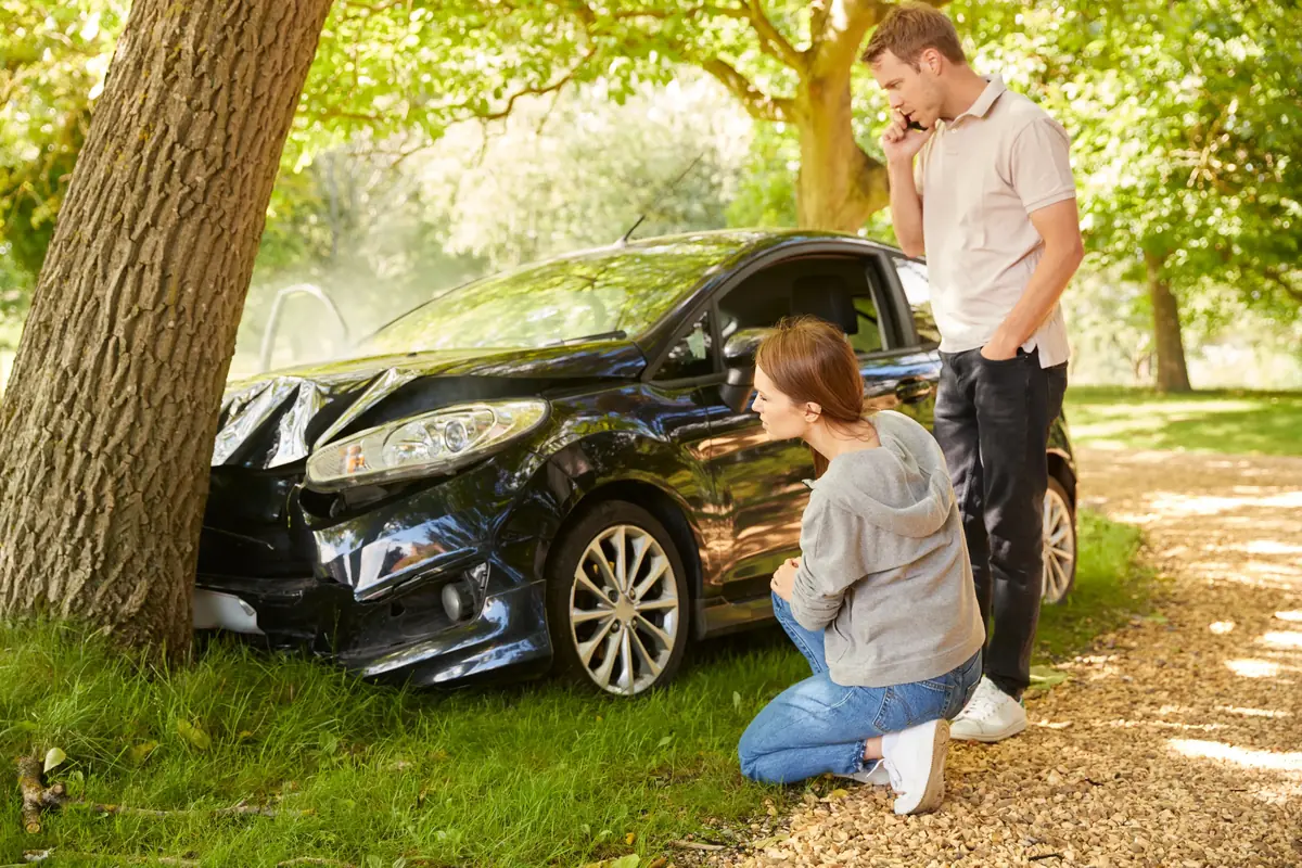 A man and woman examining a damaged car that has crashed into a tree, with the man talking on a smartphone, in a sunny park setting.