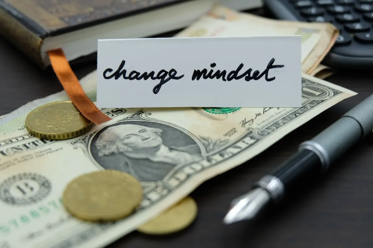 A pen, coins, and U.S. dollar bills surrounding a card with the handwritten message change mindset.