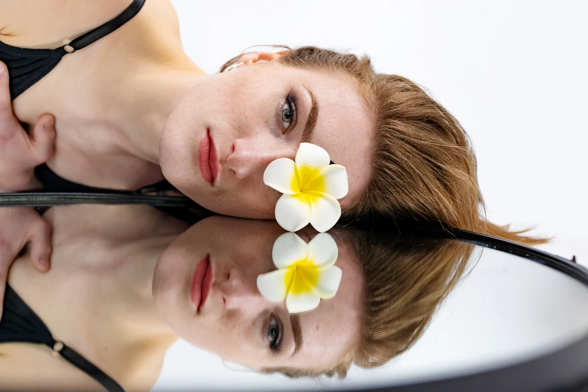 A woman rests her head on a reflective surface with a flower placed on her forehead.