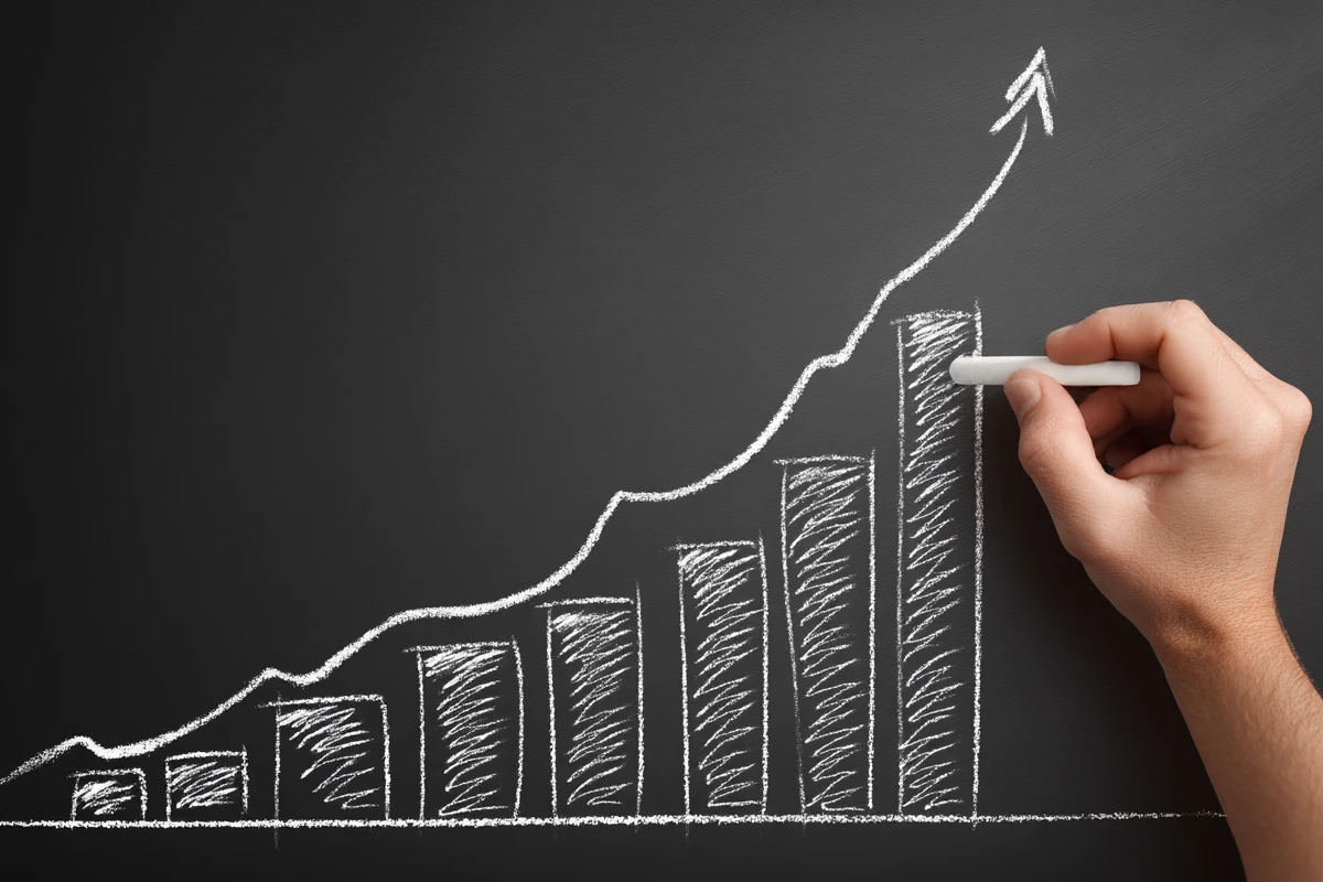 A hand draws an upward-trending bar chart with chalk on a blackboard, indicating growth or progress.