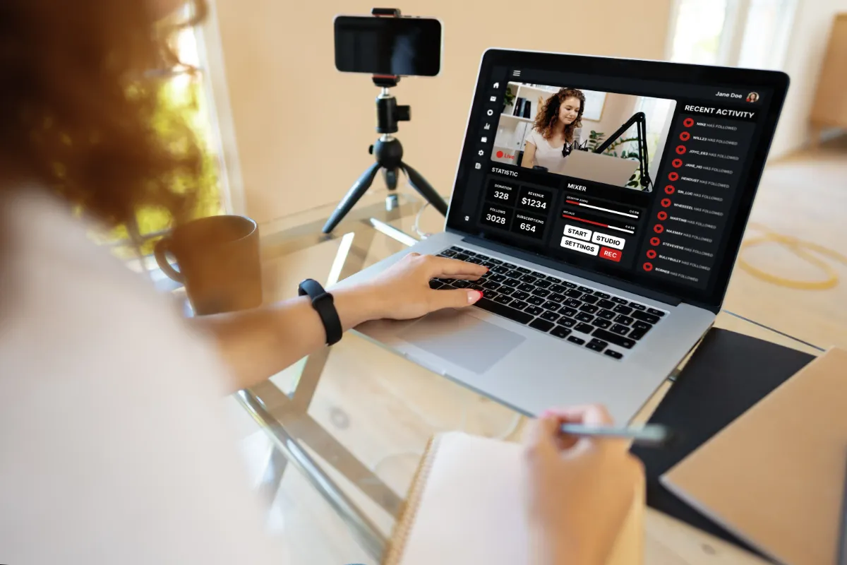 A person sitting at a desk using a laptop for video conferencing, with a smartphone on a tripod and a notebook nearby.
