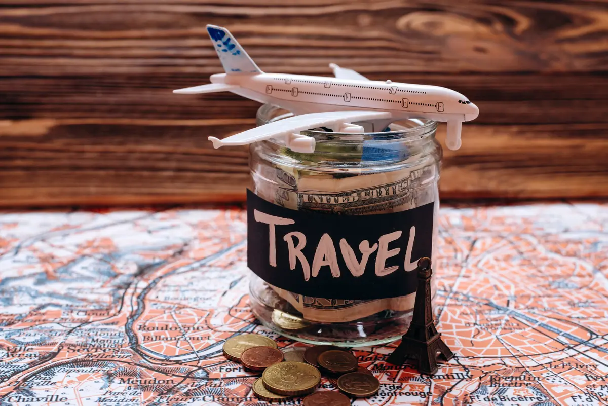 Jar labeled "Travel" filled with money, topped with a toy airplane, surrounded by coins and a small Eiffel Tower statue on a map background.