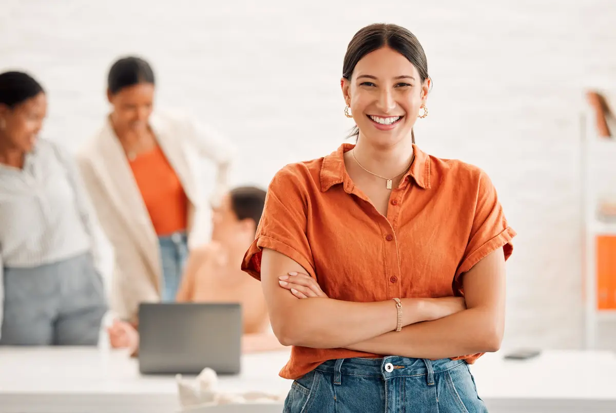 A woman in an orange shirt stands confidently with arms crossed, smiling, while a group of people engages in conversation in the background.