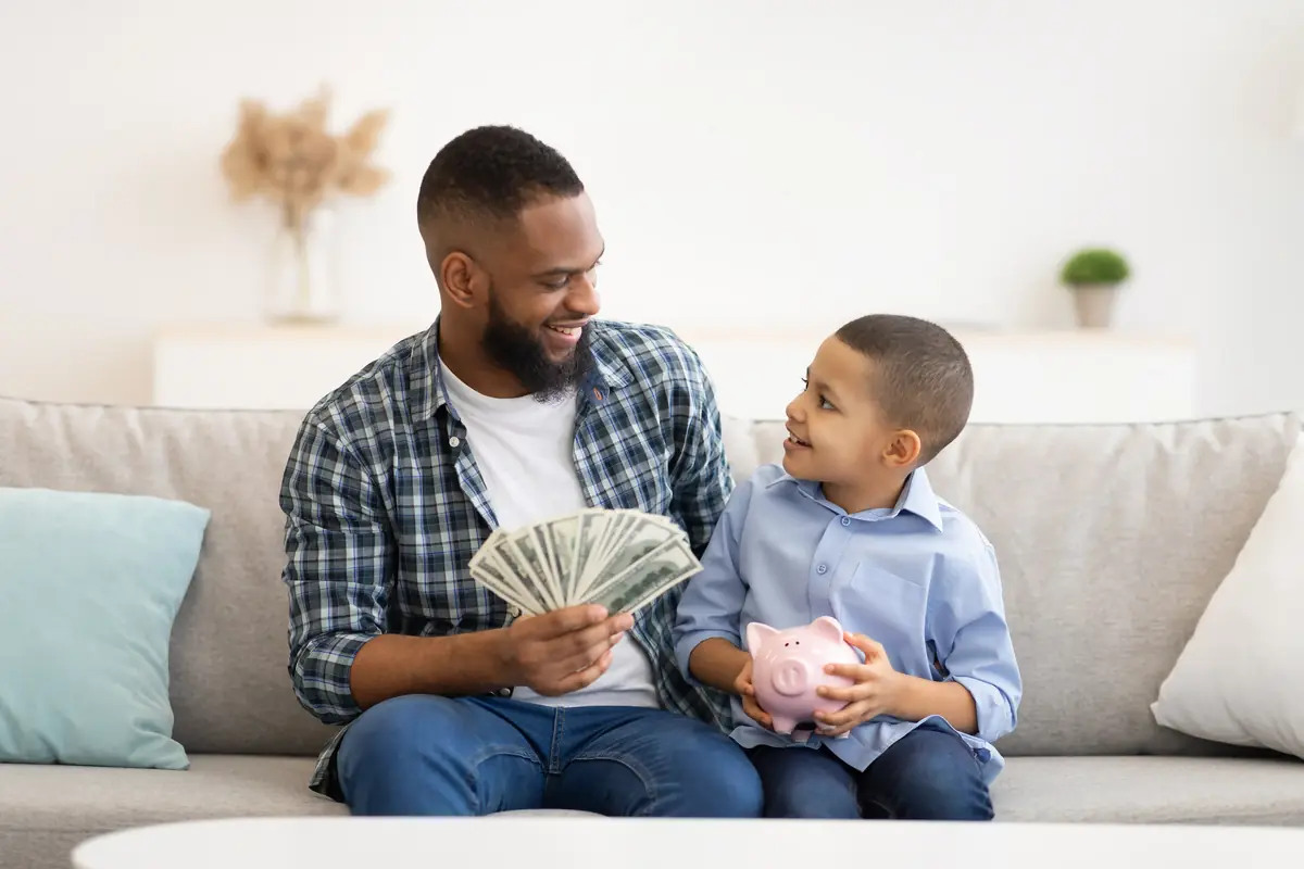 Father and son sitting on a couch, with the father holding a stack of dollar bills and the son holding a pink piggy bank, both smiling at each other.