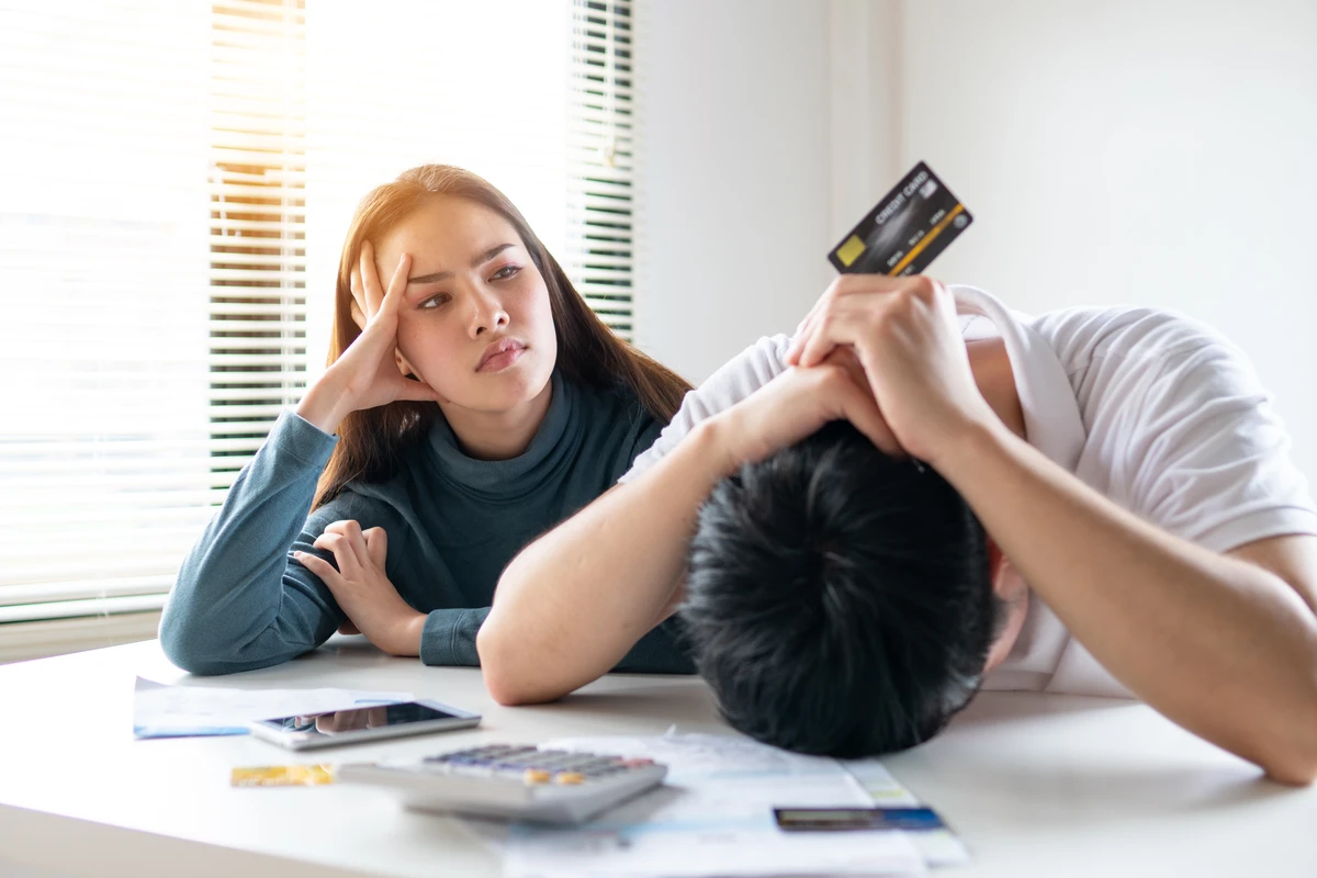 A frustrated couple sits at a table covered with financial documents, with the man holding a credit card and leaning his head on his arms, while the woman looks on with concern.
