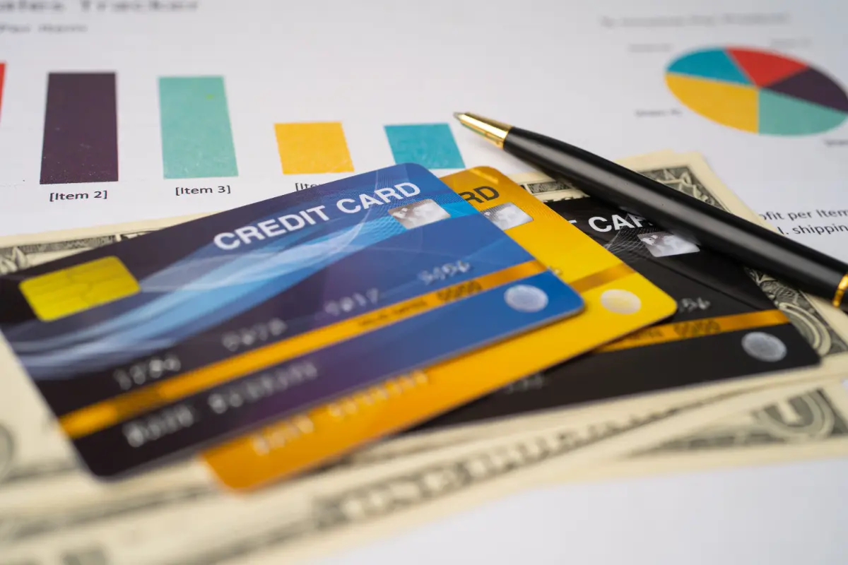 Several colorful credit cards stacked on top of one another, placed over a few U.S. dollar bills, with a pen and financial charts in the background.