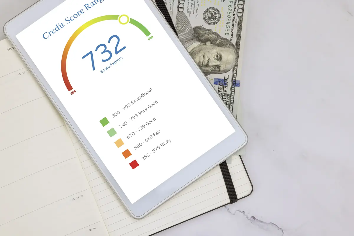 Tablet displaying a credit score of 732 with a color-coded rating scale, placed on an open notebook with hundred-dollar bills partially visible underneath.