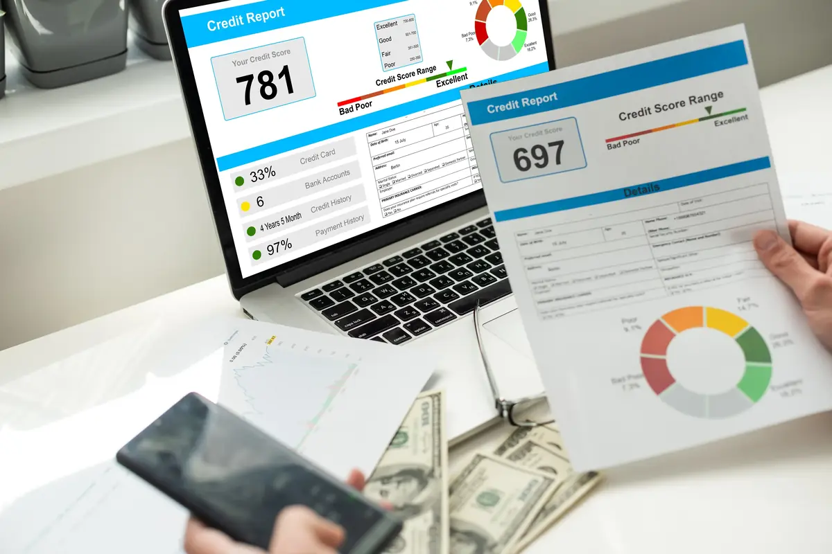 A person reviews a printed credit report with a score of 697, while a laptop displays another report with a score of 781, surrounded by financial documents and dollar bills.