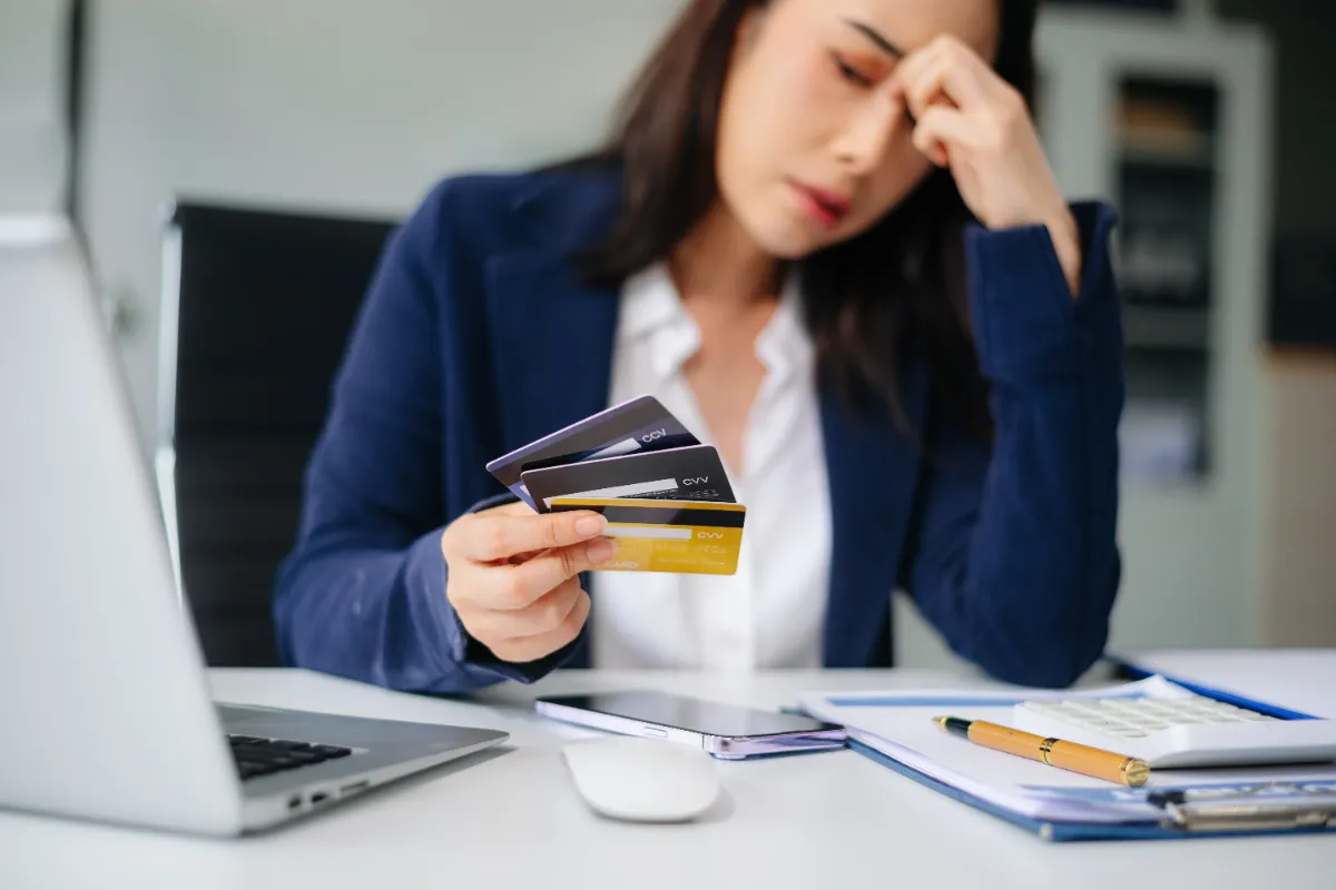 A woman in a blazer holding multiple credit cards, sitting at a desk with a laptop and documents, looking stressed.