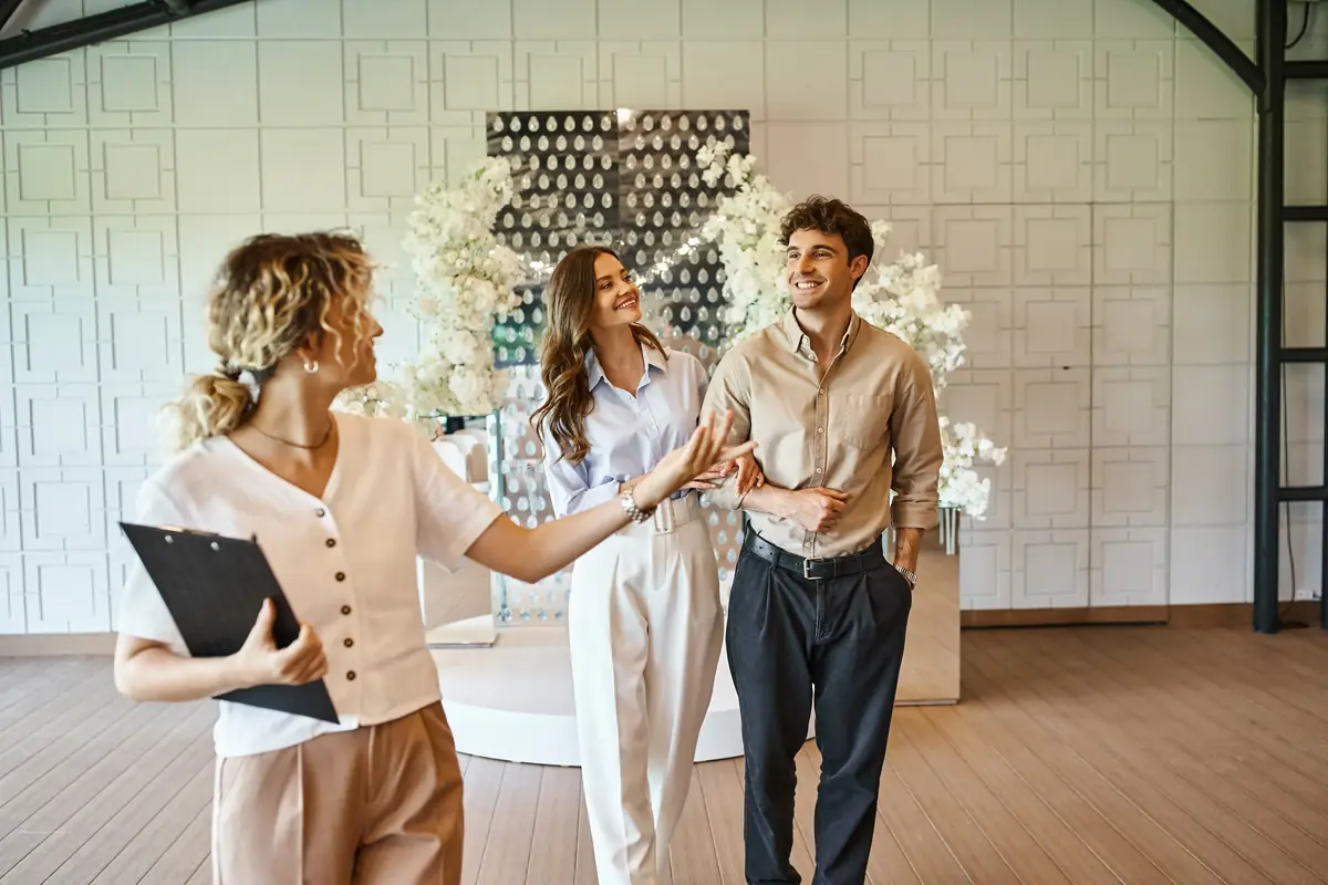 A woman with a clipboard guides a smiling couple through a decorated venue, featuring white floral arrangements and a modern backdrop.