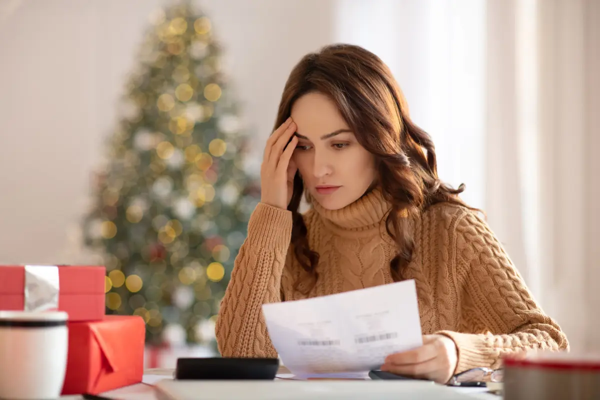 Woman in a cozy sweater looks concerned while reviewing documents at a table, with wrapped gifts and a decorated Christmas tree in the background.