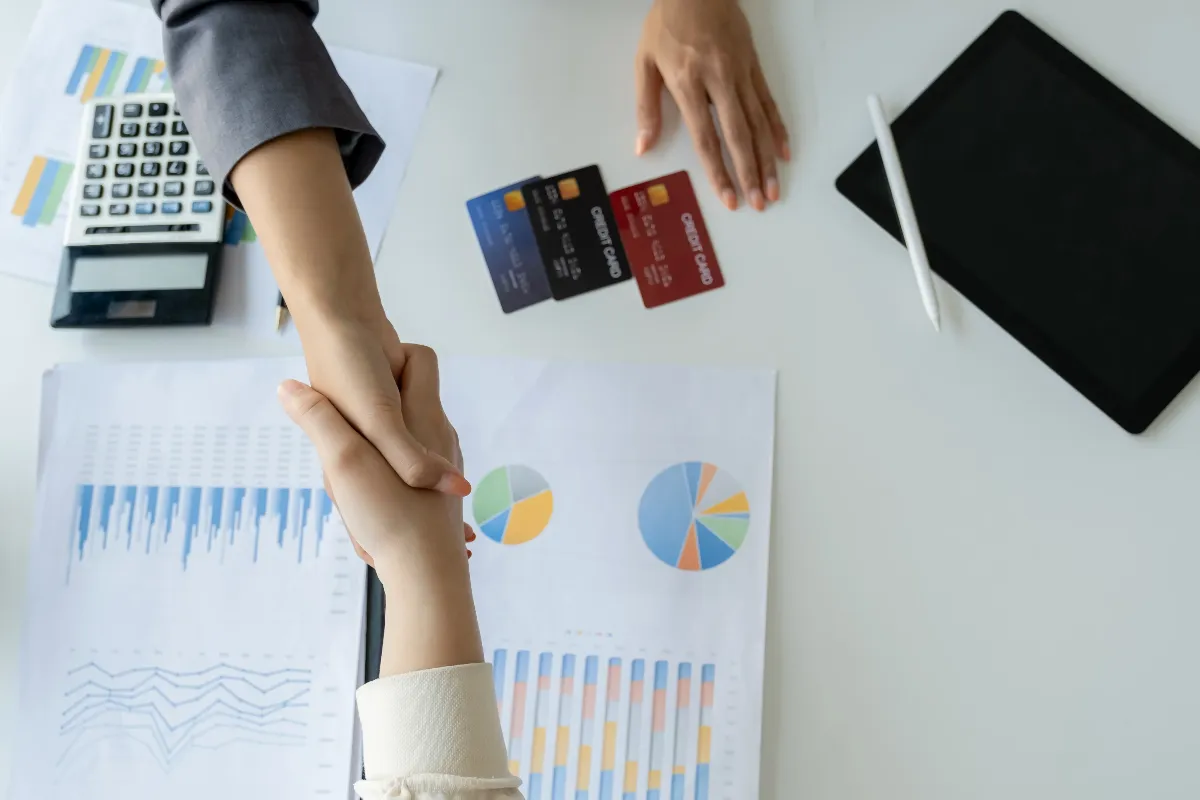 Two people shaking hands over a desk with credit cards, financial charts, a calculator, and a tablet, suggesting an agreement on financial or debt consolidation.
