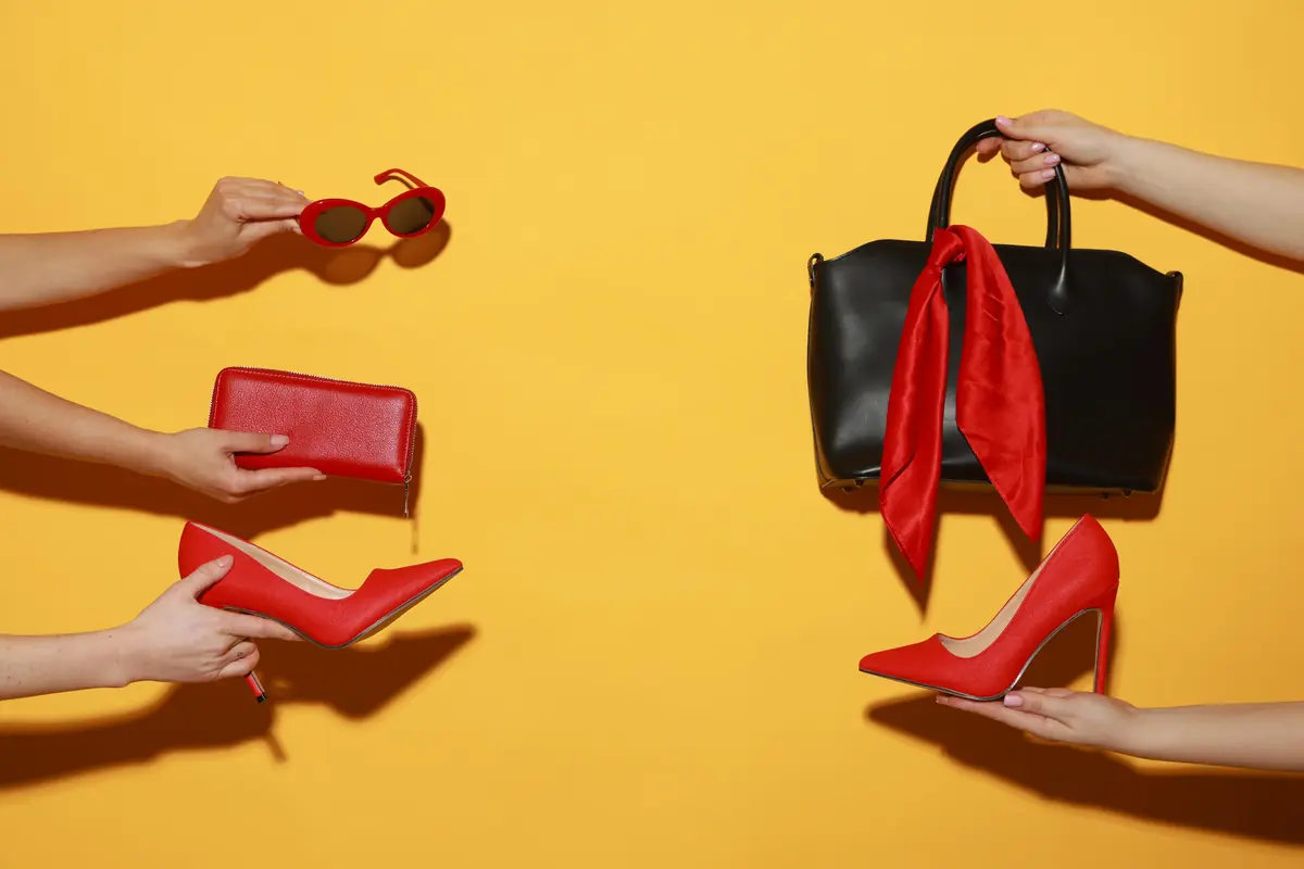 Four hands display various fashion accessories against a yellow background, including black sunglasses, a black handbag with a red scarf, a red wallet, and red high-heeled shoes.